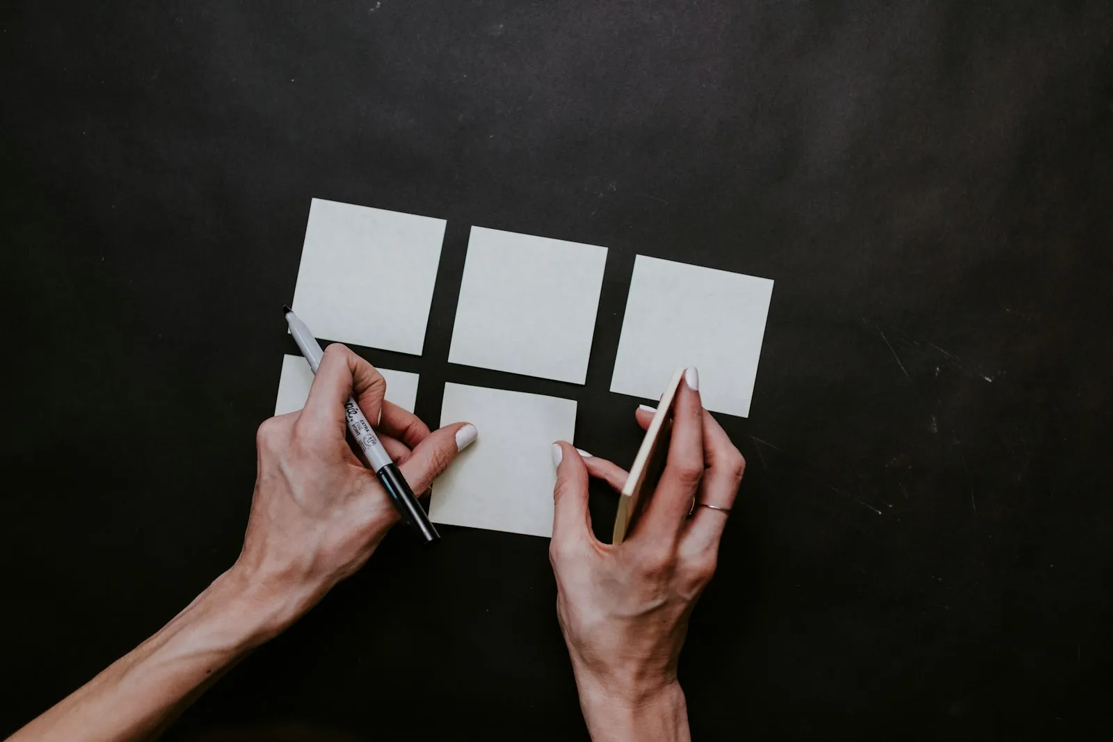 Mãos sobre uma mesa escura organizando post-its em branco, uma delas segurando uma caneta